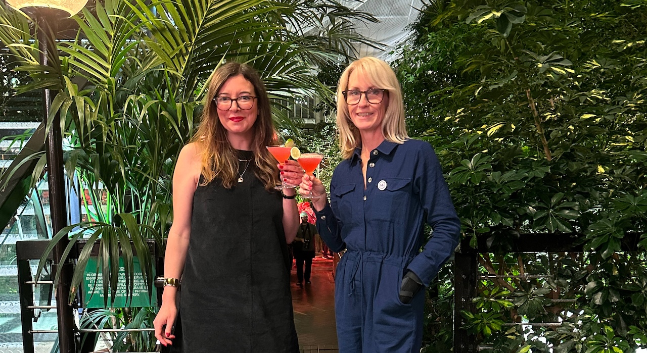 Two smartly dressed women holding cocktails in front of some foliage.